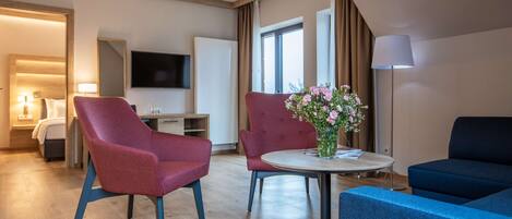 A modern hotel room with a wooden floor, a round dining table with chairs, a sofa, a TV, and a lamp.