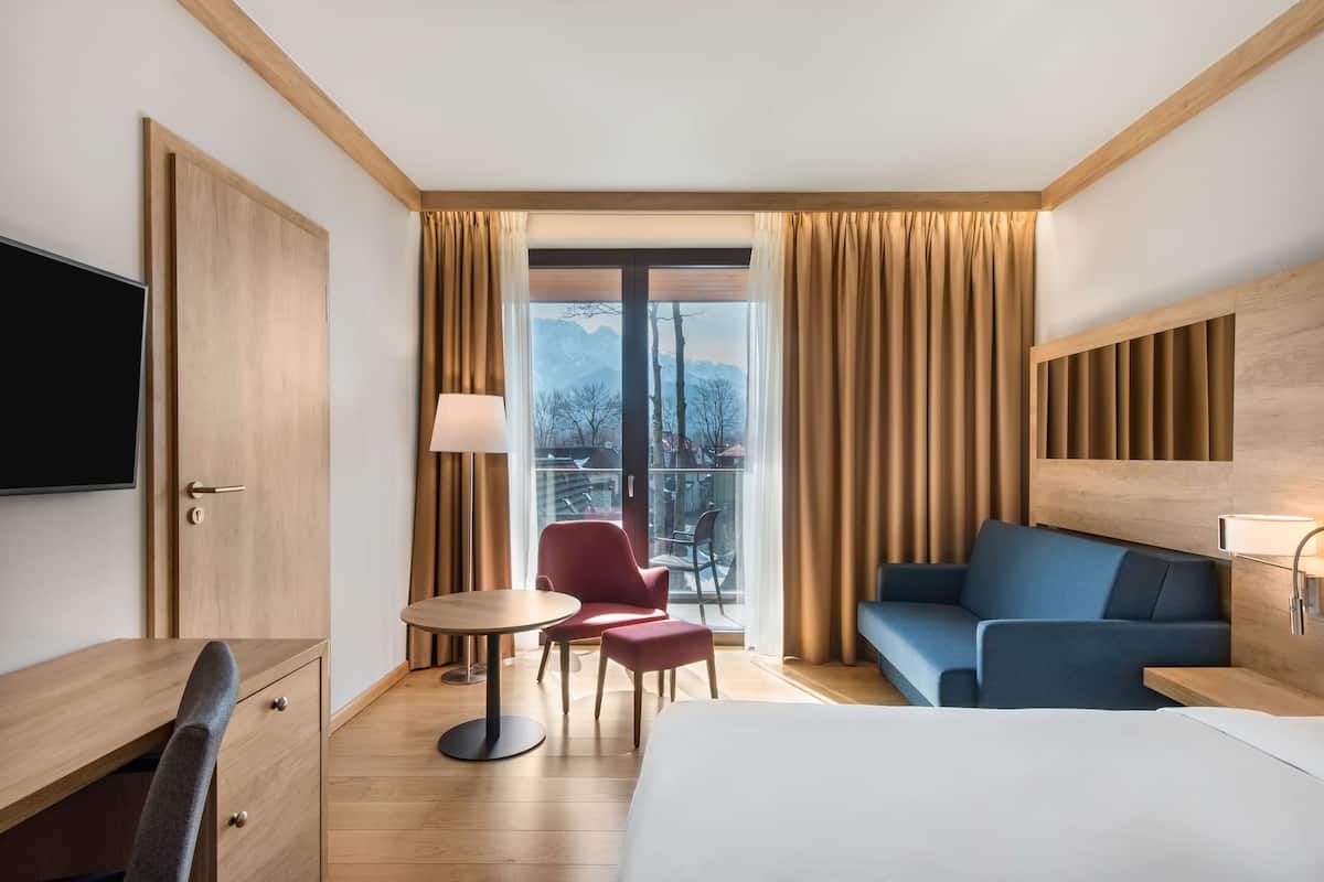 Habitación de hotel con cama, escritorio, silla, sofá, televisor y balcón con cortinas.