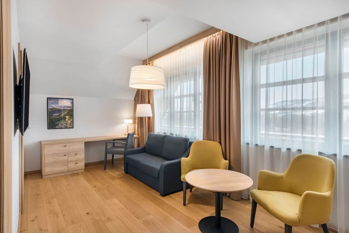 Una habitación de hotel moderna con suelo de madera, un sofá, un sillón amarillo, una mesa redonda, un escritorio con silla, una lámpara, un televisor y una ventana con cortinas.