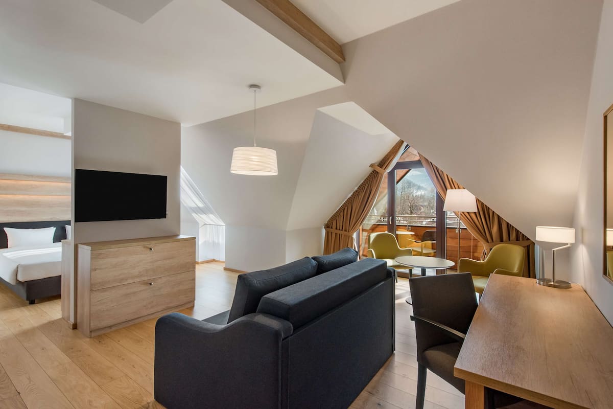Habitación de hotel moderna con sofá, televisión, mesa de comedor y un pequeño balcón con vistas.