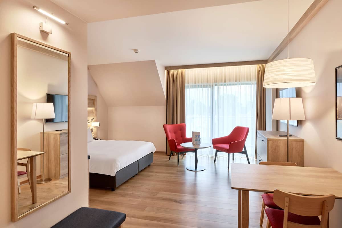 Una habitación de hotel con una cama, un escritorio, dos sillas rojas, una mesa redonda y un espejo grande.