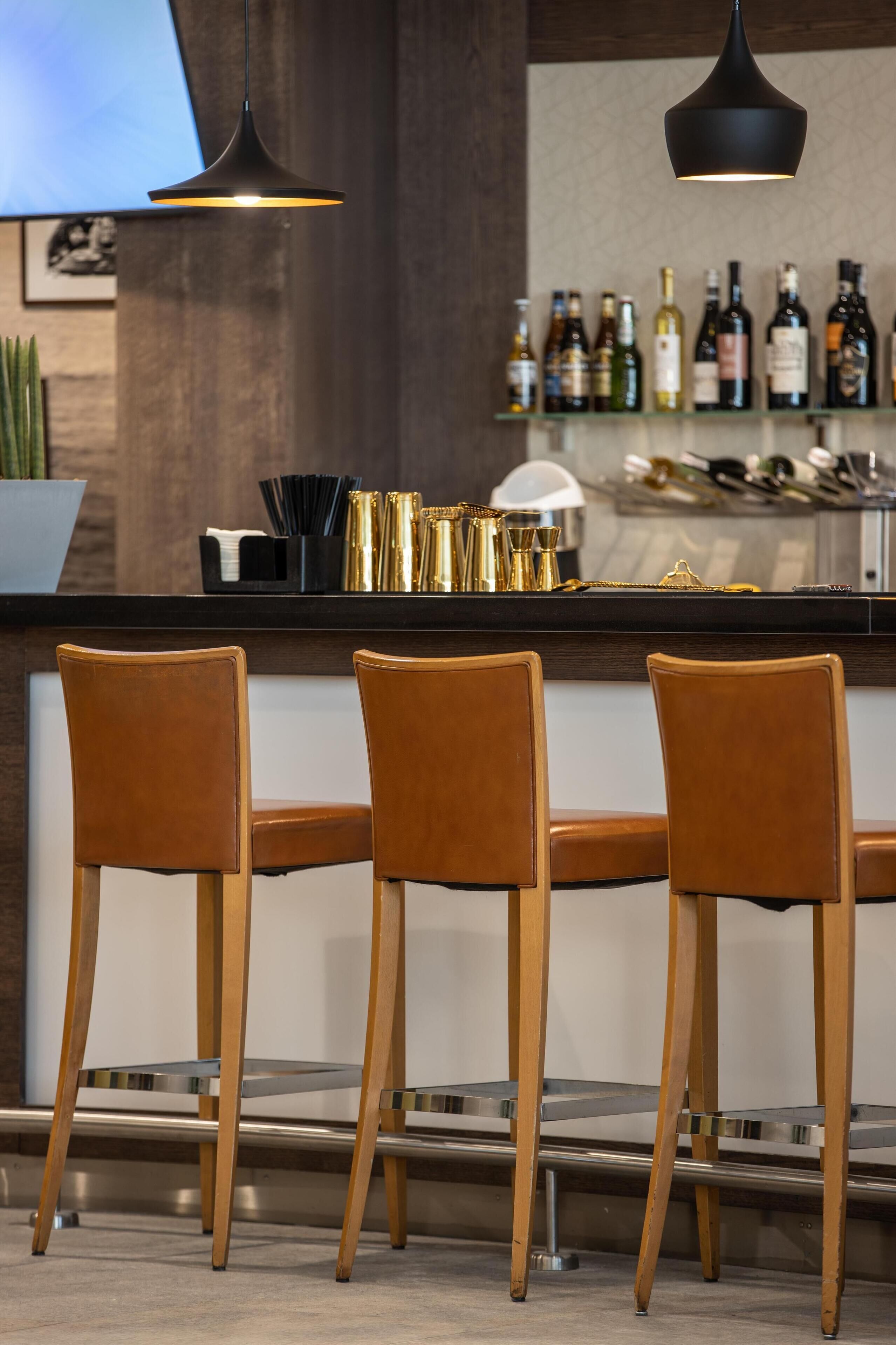 A bar area with three brown leather bar stools, a black pendant light, and a shelf with various bottles.