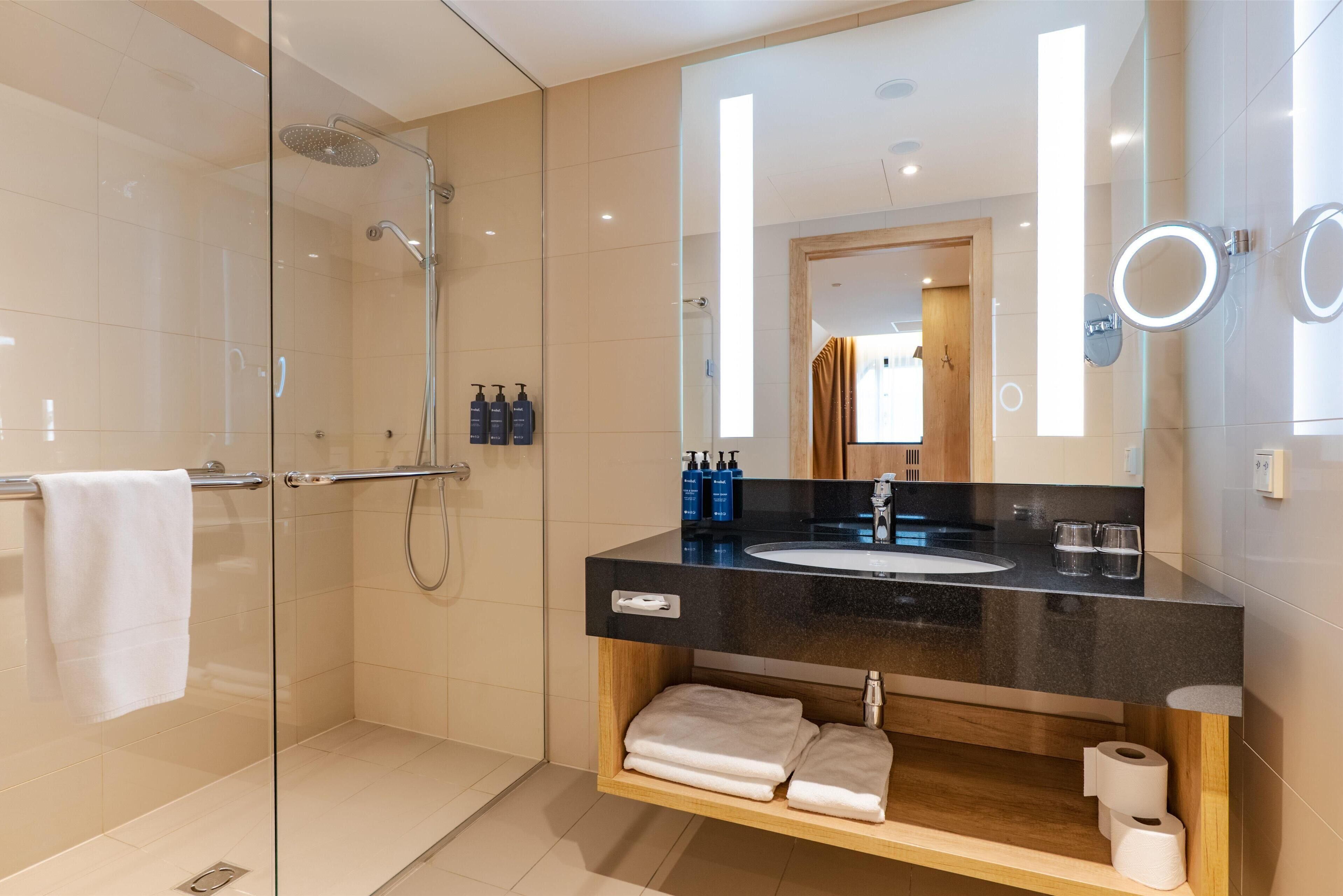 A modern bathroom with a glass-enclosed shower, a black countertop sink, wooden vanity, and a large mirror with integrated lighting.