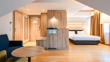 Una habitación de hotel moderna con cama de altillo, una pequeña kitchenette, una zona de estar con una mesa redonda y un amplio ventanal con cortinas.