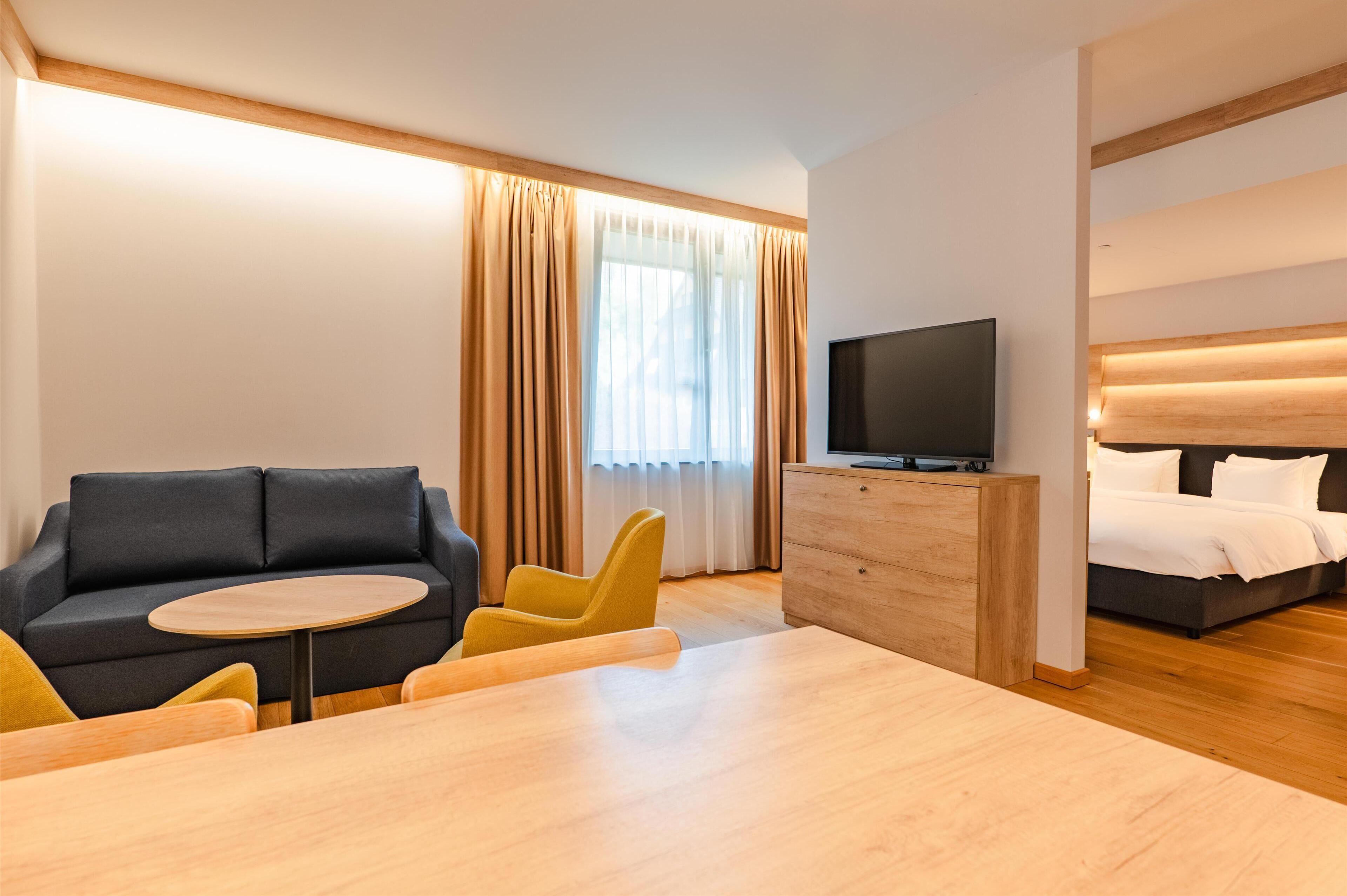 A modern hotel room with a sofa, a small round table, a TV, a bed, and a wooden dresser.