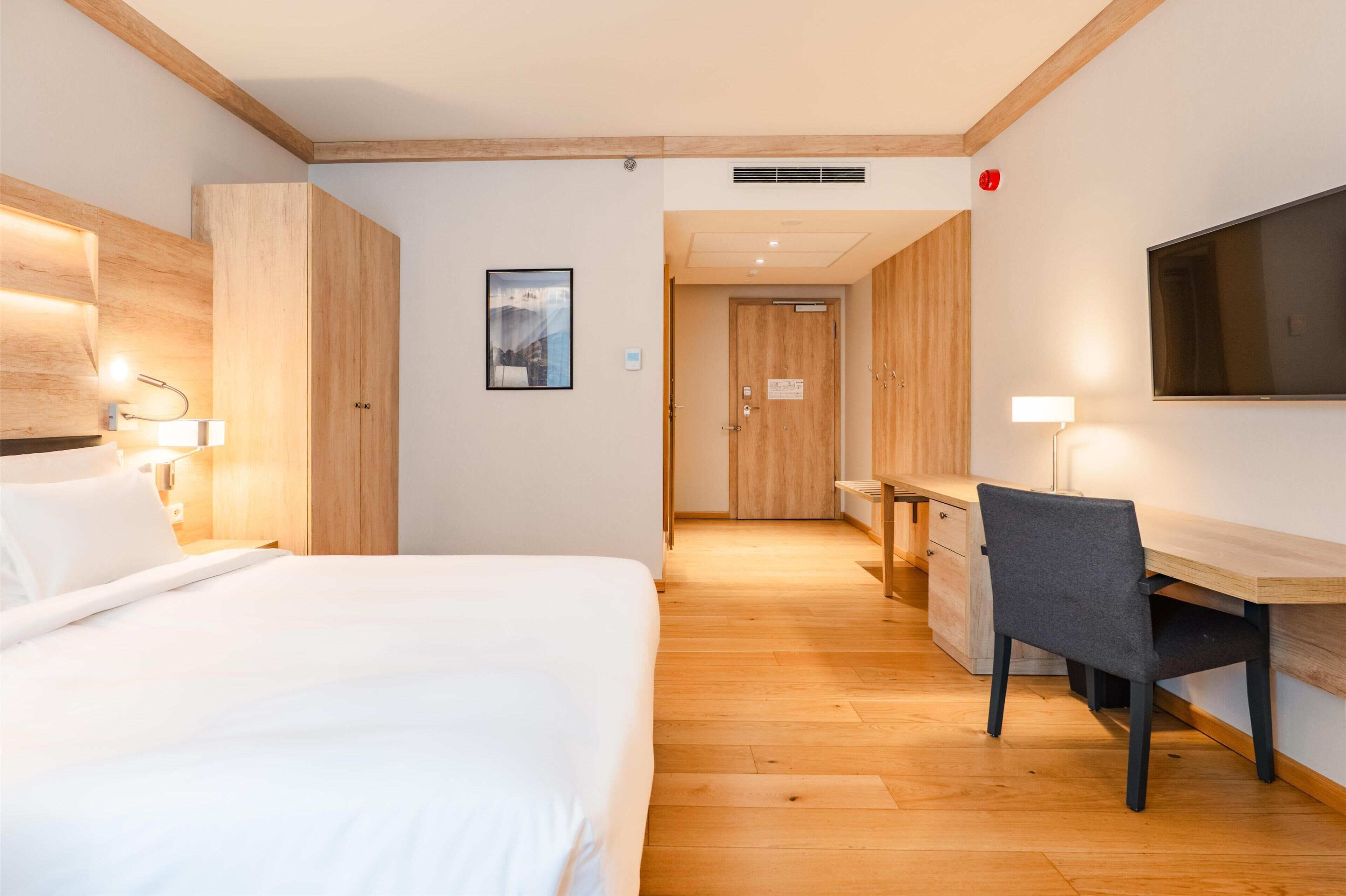 A hotel room with a bed, desk, chair, and a TV mounted on the wall.