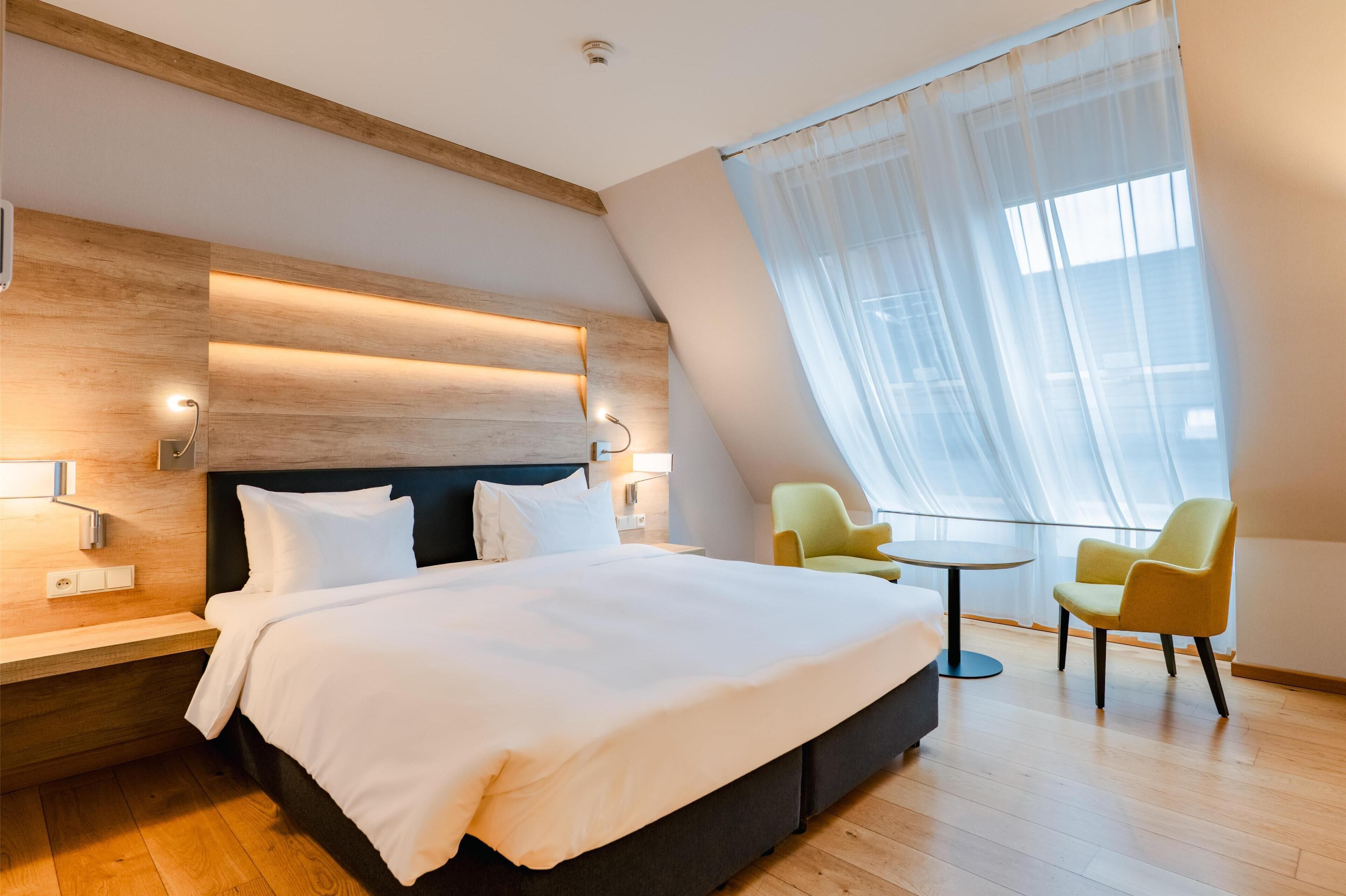 Ein modernes Hotelzimmer mit einem großen Bett, einem hölzernen Kopfteil, einem kleinen runden Tisch, zwei Stühlen und einem großen Fenster mit transparenten Vorhängen.