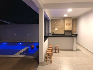 A modern indoor pool area with a bar and stools, a tiled floor, and a counter with a sign. - Casa com piscina em Caraguatatuba (Caraguatatuba)