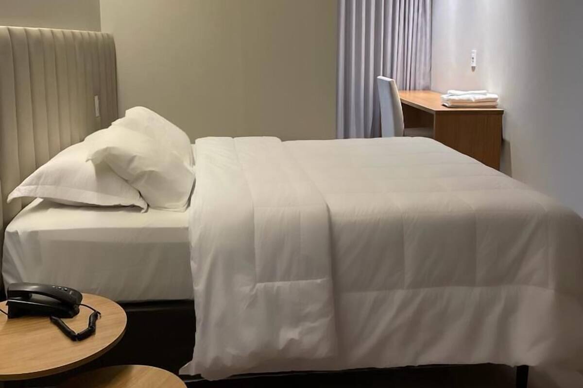 A hotel room with a bed, bedside table, chair, desk, and an air conditioning unit.