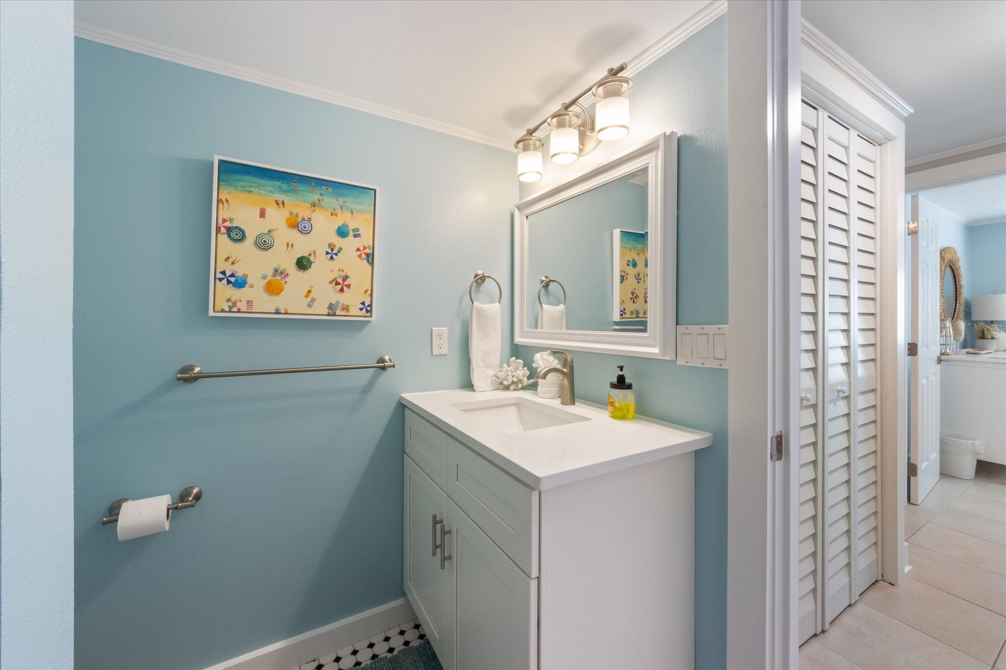 A bathroom with a light blue wall, a sink with a mirror above it, a towel rack, and a toilet paper holder.