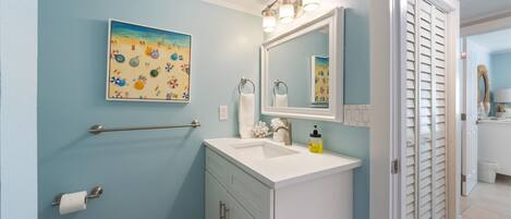 A bathroom with a light blue wall, a sink with a mirror above it, a towel rack, and a toilet paper holder.