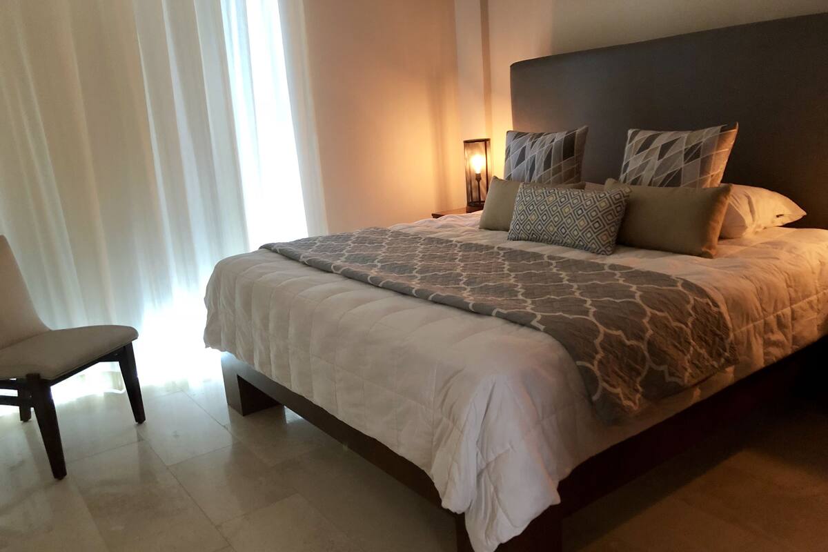 A neatly made bed with patterned bedding and pillows, a bedside table with a lamp, and a chair in a well-lit room with a large window.