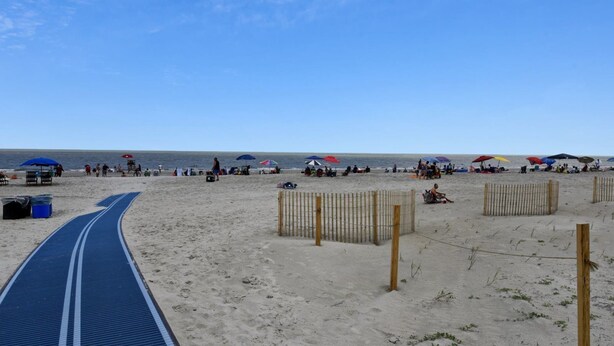 Une plage avec un chemin bleu menant à l’eau, des clôtures en bois et des parasols de plage.