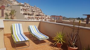 Een terras op het dak met twee gestreepte lounge stoelen, potplanten en uitzicht op een woongebied met terrasvormige huizen.