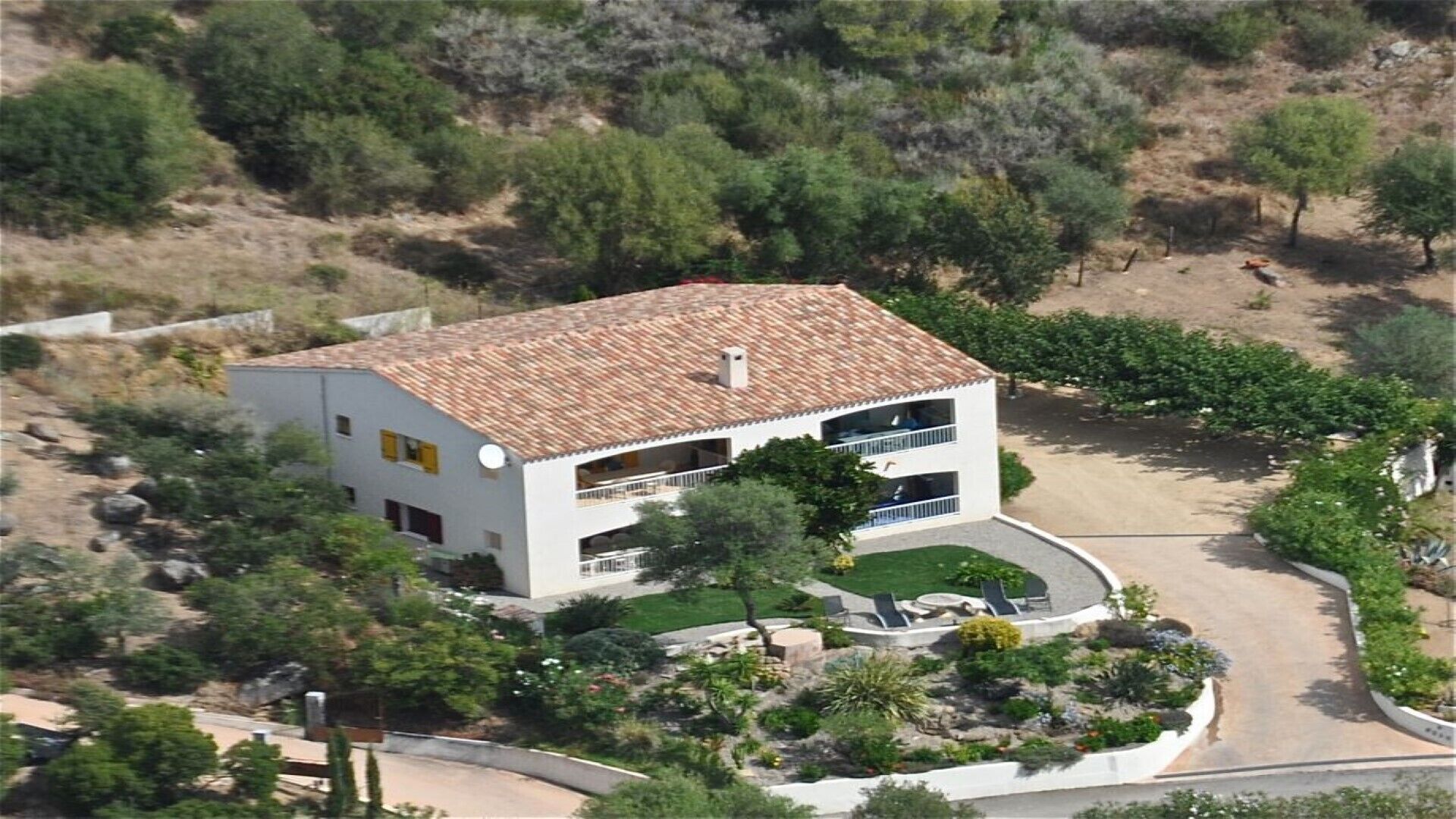 Une maison à deux étages, avec un toit recouvert de tuiles, entourée d’un jardin aménagé où poussent différentes espèces de plantes, et dotée d’une allée.