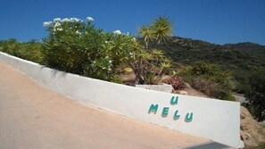Paredes brancas com o texto "U MELU" em letras verdes, rodeadas por vegetação e um céu azul claro.