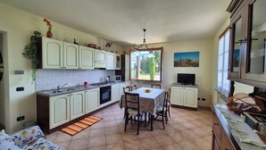 Una cucina con mobili bianchi, un tavolo da pranzo con sedie, una televisione e una finestra vista alberi.