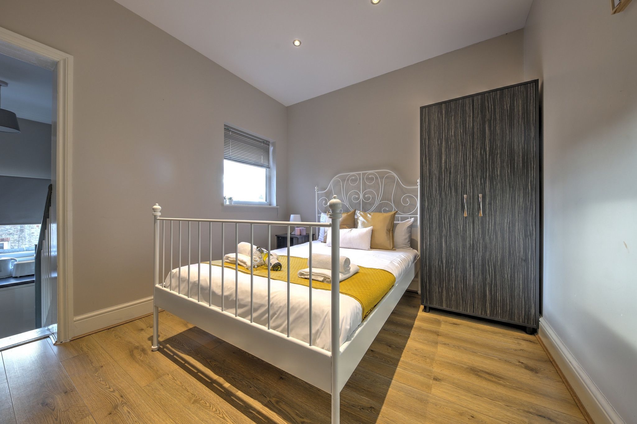 A bedroom with a white metal bed frame, a wooden floor, a large wardrobe, and a window with blinds.