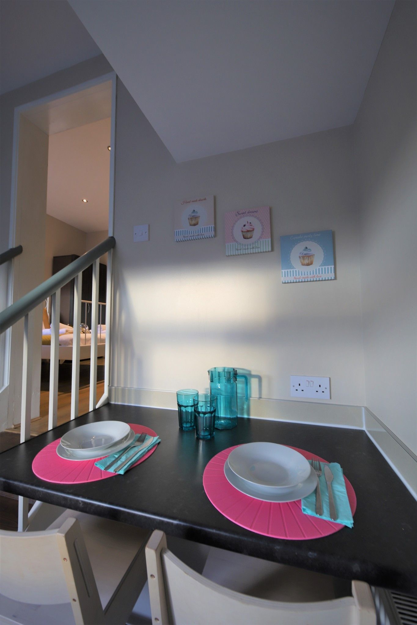 Un comedor con una mesa negra, individuales rosa y una pared blanca decorada con motivos de cupcakes.
