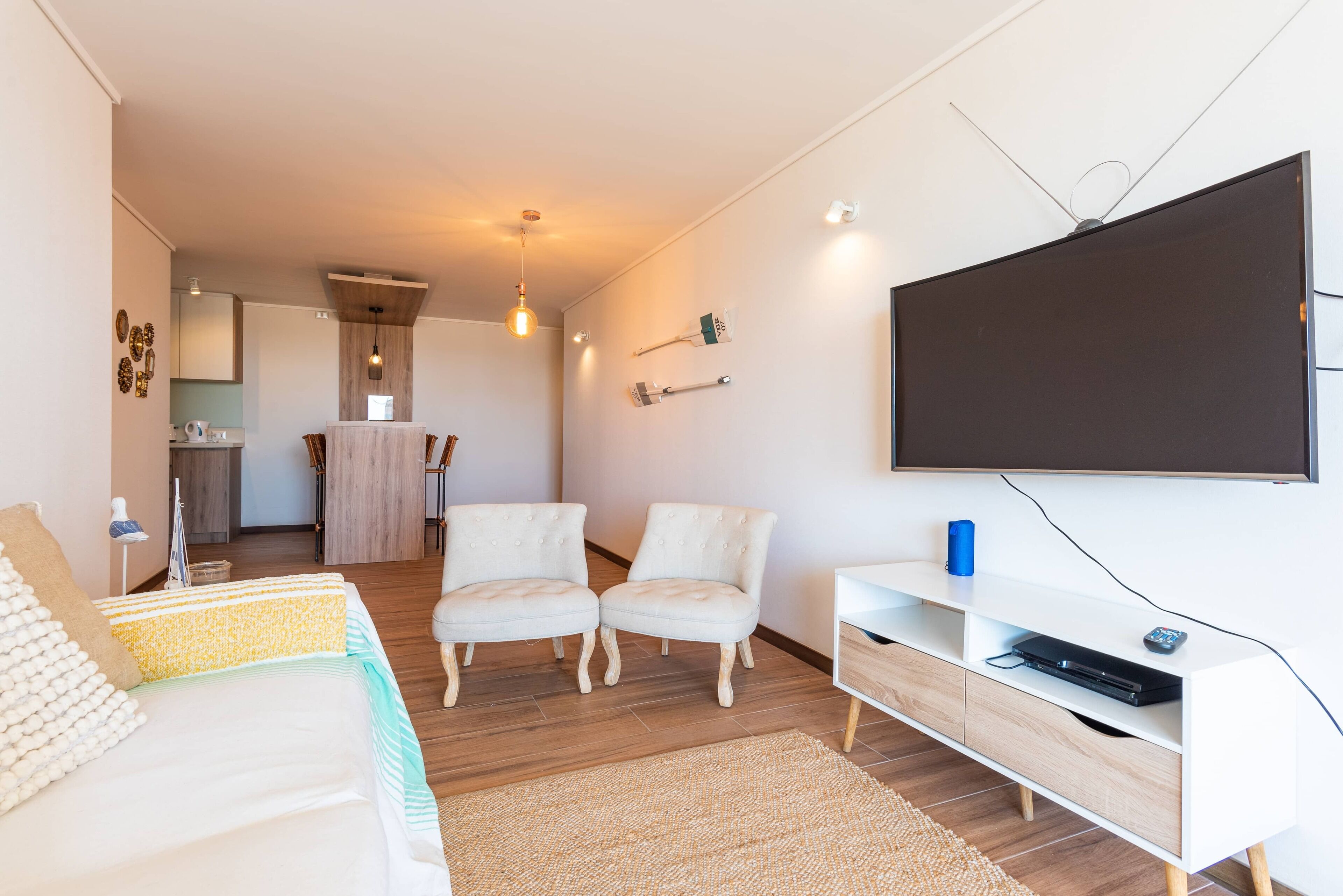 A modern hotel room with a bed, two chairs, a TV, and a small kitchenette in the background.
