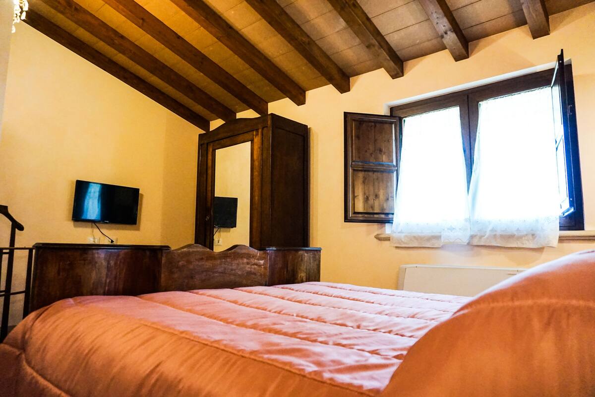 Une chambre avec un plafond en bois, un lit recouvert d’une couverture rose, une commode en bois, un téléviseur à écran plat et une fenêtre avec des rideaux.