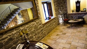 A stone-walled room with a staircase, a small window with a view, a table with a clock, and a chair.