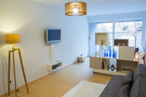 Ein modernes Wohnzimmer mit Flachbildfernseher, Stehlampe, Sofa und einem Fenster mit Blick auf Bäume.
