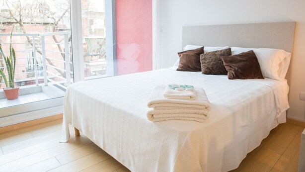 A neatly made bed with a white bedspread and pillows, a stack of folded towels, and a potted plant on the windowsill. - Borges Apartment by Be Local Argentina (Buenos Aires)