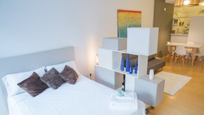 A hotel room with a bed, a white headboard, a white nightstand, a white dresser, a small table with a chair, and a colorful painting on the wall. - Borges Apartment by Be Local Argentina (Buenos Aires)
