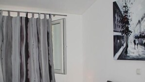 Ein Schlafzimmer mit einem Bett, Kissen, einem Nachttisch, einer Lampe und einem Fenster mit Vorhängen.