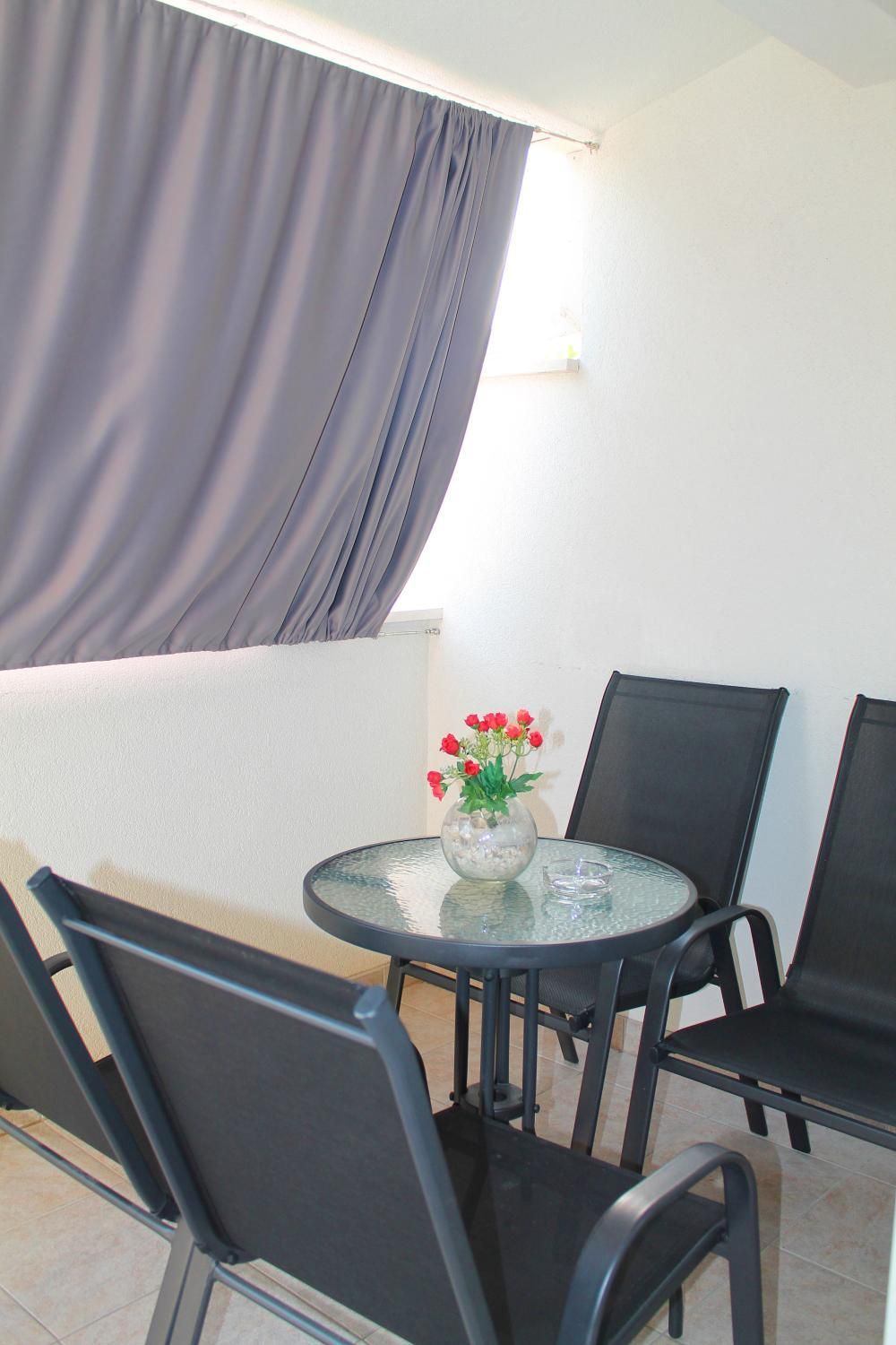 A small round table with chairs, a vase with red flowers, and a window with curtains.