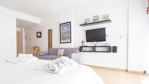 Moderni hotellihuone, jossa on suuri sänky, harmaa sohva, seinälle asennettu litteänäyttöinen televisio ja pieni sivupöytä.