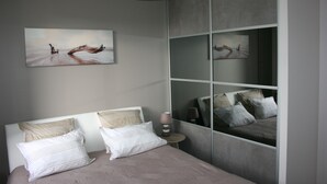 Une chambre à coucher moderne avec un lit, des oreillers, une table de chevet et un miroir fixé au mur qui reflète la pièce.