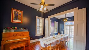 Quarto com um piano, uma mesa de jantar com cadeiras, um ventilador de teto e uma porta que dá para outro espaço.