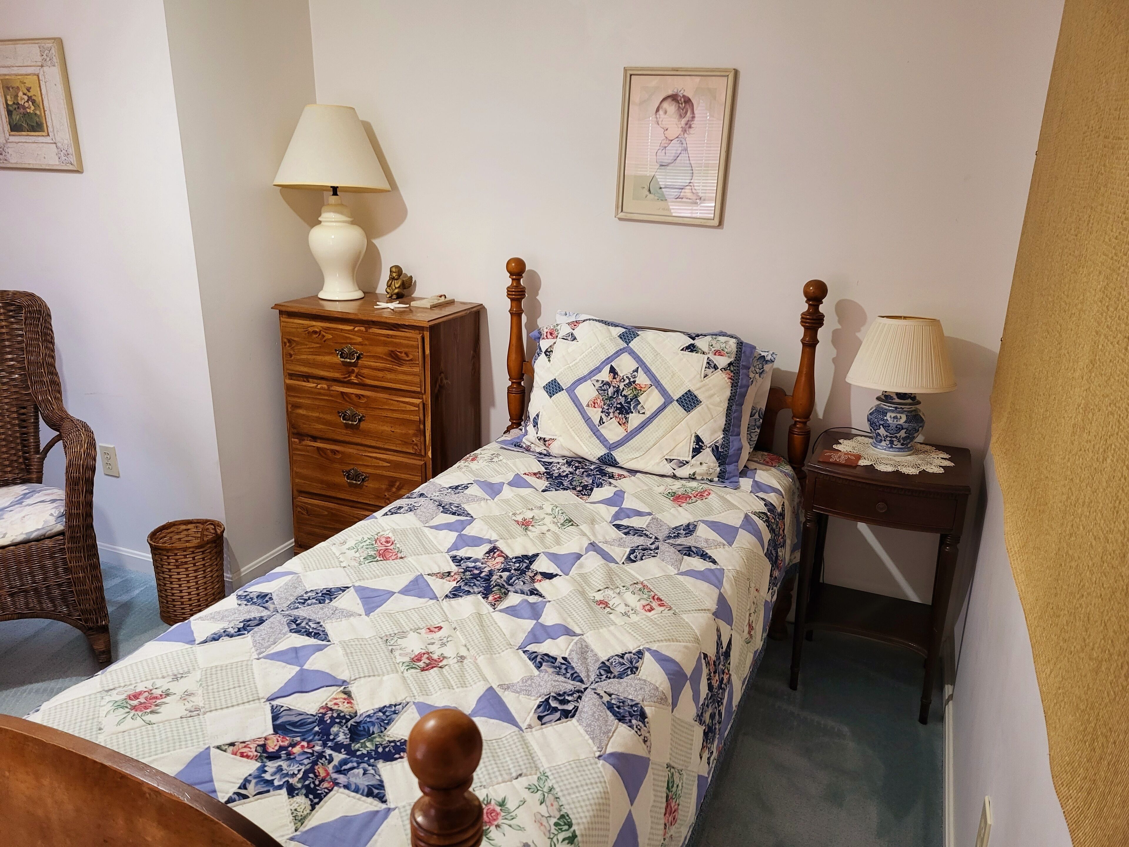 Un dormitorio con cama, cómoda, mesita de noche, lámpara y silla de mimbre.