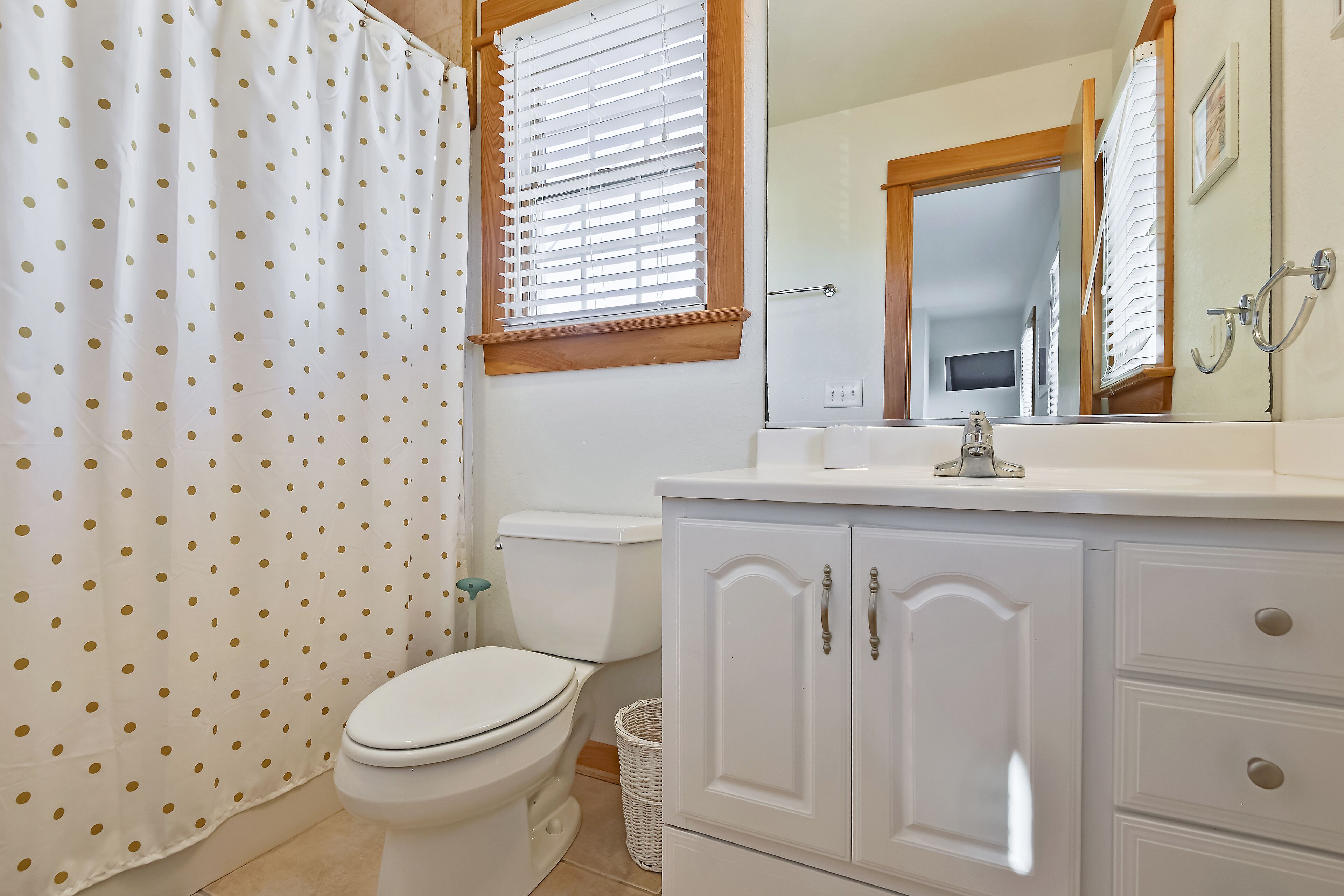 A bathroom with a toilet, a white cabinet with drawers, a shower curtain with gold polka dots, and a window with blinds.