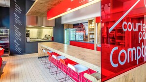 A modern café interior with a red and white color scheme, featuring a long counter with stools, a visible kitchen area, and a chalkboard menu. - ibis budget Limoges (Limoges)