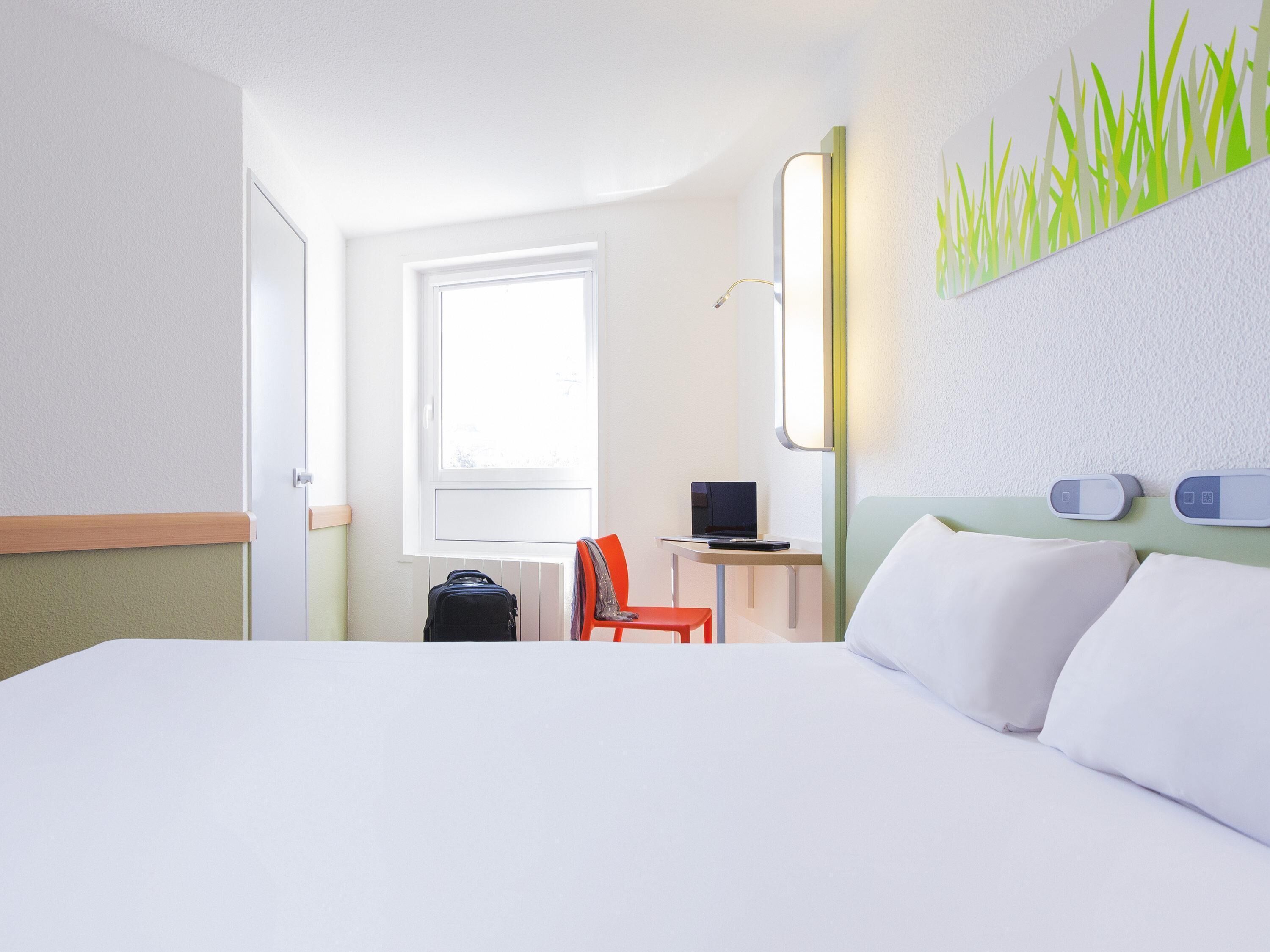 Quarto de hotel com uma cama, uma escrivaninha com um laptop, uma cadeira, uma mala e uma janela.