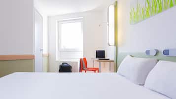 Quarto de hotel com uma cama, uma escrivaninha com um laptop, uma cadeira, uma mala e uma janela.