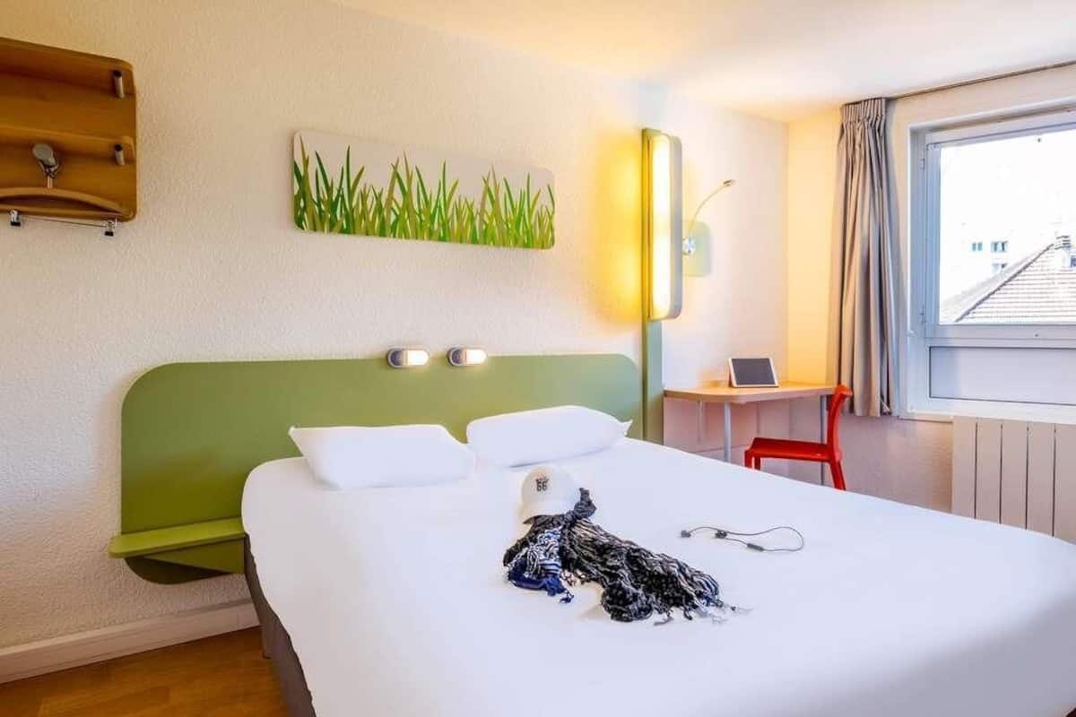 A hotel room with a green headboard, a bed with white linens, a desk with a chair, and a window with curtains.