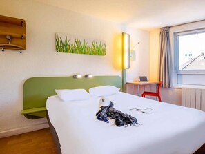 Habitación de hotel con cabecera verde, cama con ropa blanca, escritorio con silla y ventana con cortinas.