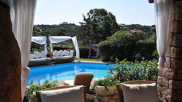 Relax by the serene pool surrounded by lush greenery and a stone wall, with comfortable lounge chairs and a shaded canopy.