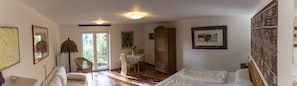 Ein Zimmer mit einem Bett, einem Esstisch mit Stühlen, einem Korbsessel, einem Schrank und einem großen Fenster.