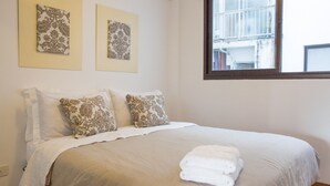 Ein ordentlich bezogenes Bett mit einer beige farbenen Tagesdecke und zwei Zierkissen, ein Fenster mit Blick auf Gebäude und zwei gerahmte Kunstwerke an der Wand.