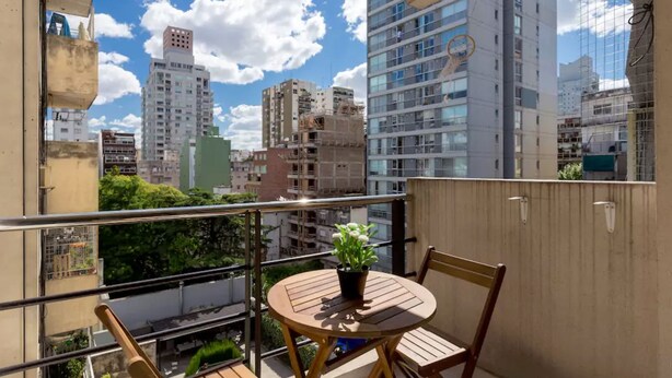 Ein Balkon mit einem runden Holztisch und Stühlen, einem Blumentopf und Blick auf hohe Gebäude.