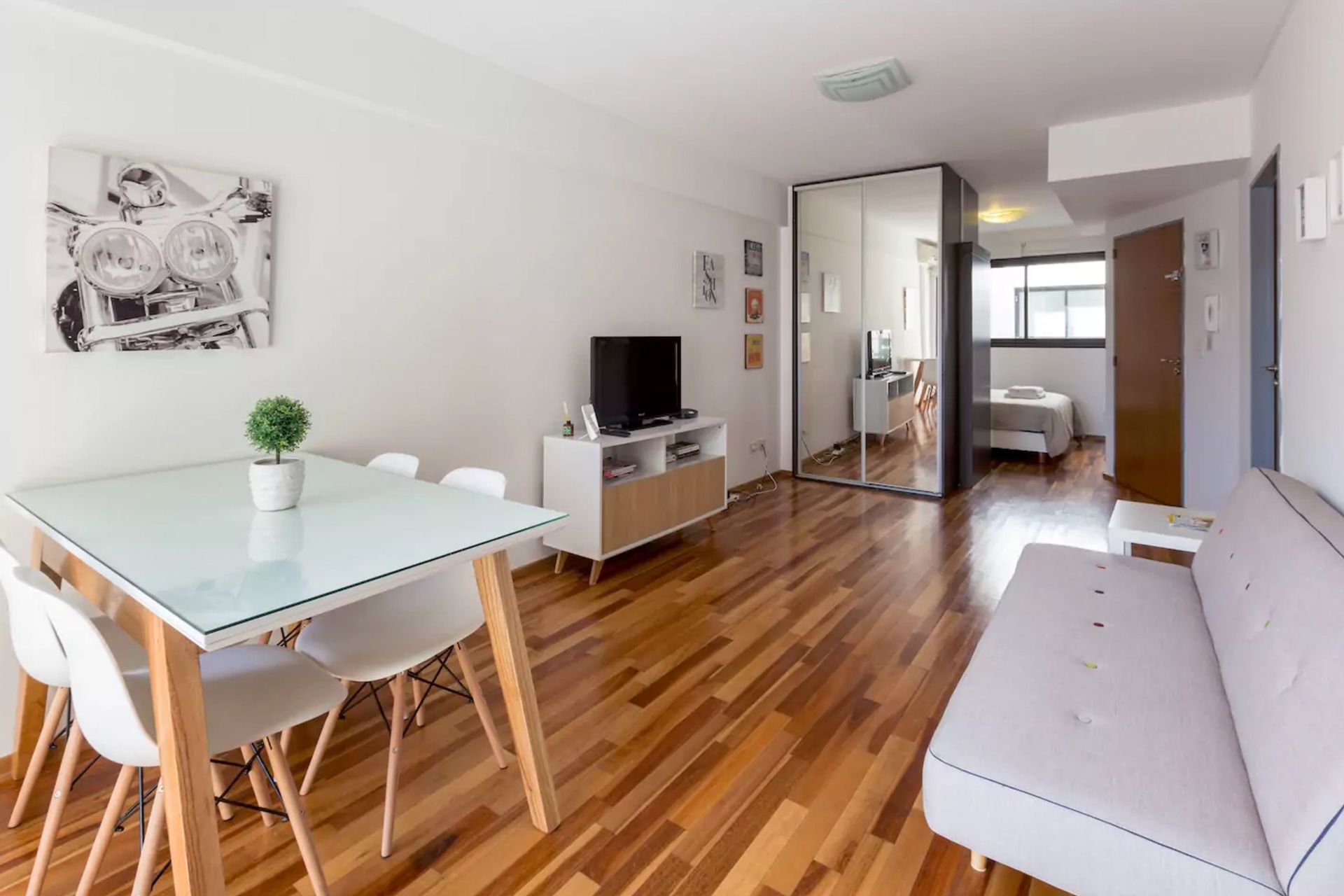 A modern living room with a glass dining table, white chairs, a white sofa, and a television on a wooden cabinet.