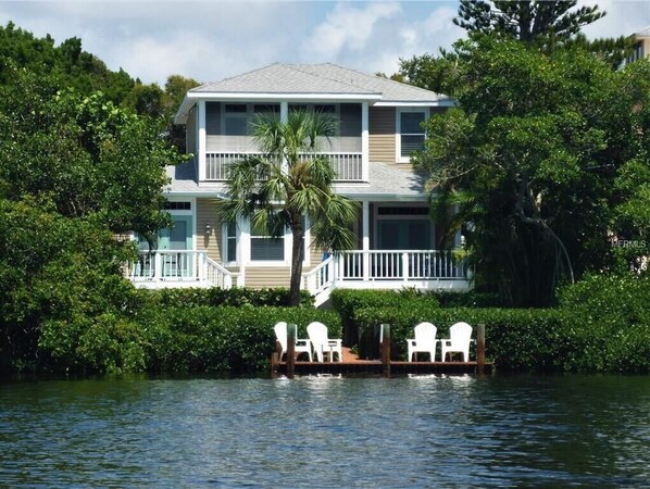 A two-story house with a balcony, surrounded by lush greenery and a dock with chairs.