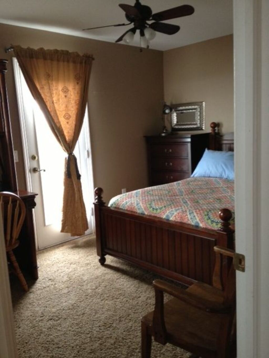Un dormitorio con cama de madera, cómoda, silla, ventilador de techo y una puerta con cortinas.