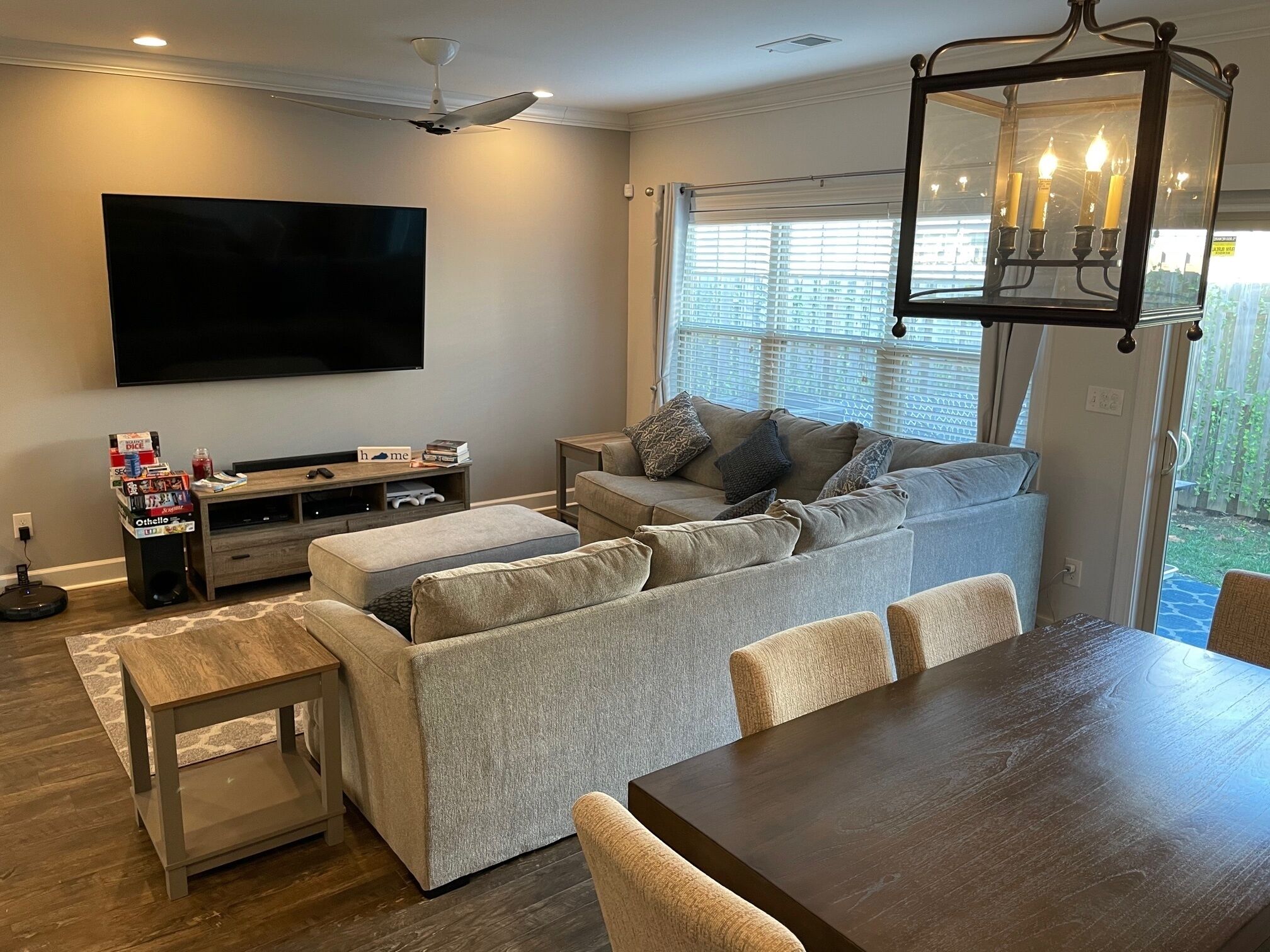 A living room with a flat-screen TV, a ceiling fan, a dining table with chairs, and a large window with blinds.