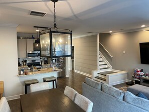 Una cocina moderna con una isla central, un comedor con mesa de madera y sillas tapizadas en gris, y una escalera que sube a un segundo piso.